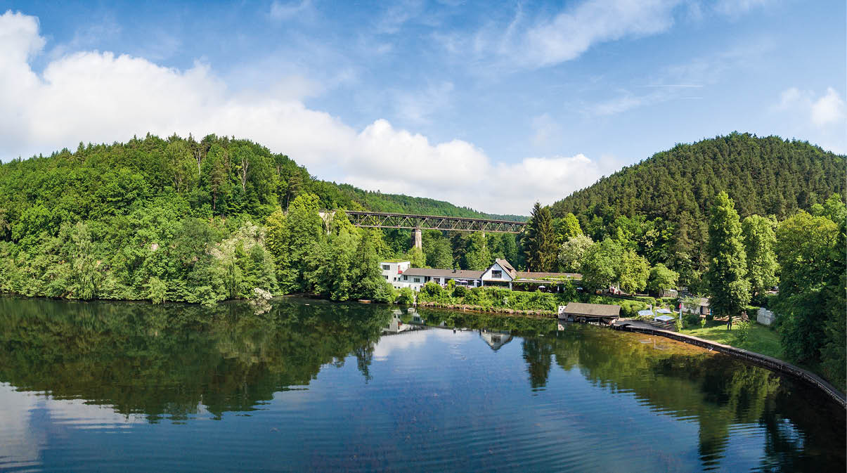 Ein ruhiger See spiegelt die umliegenden gr nen W lder und einen kleinen Ort mit H usern und einer Br cke im Hintergrund wider. Beschreibung von KI generiert