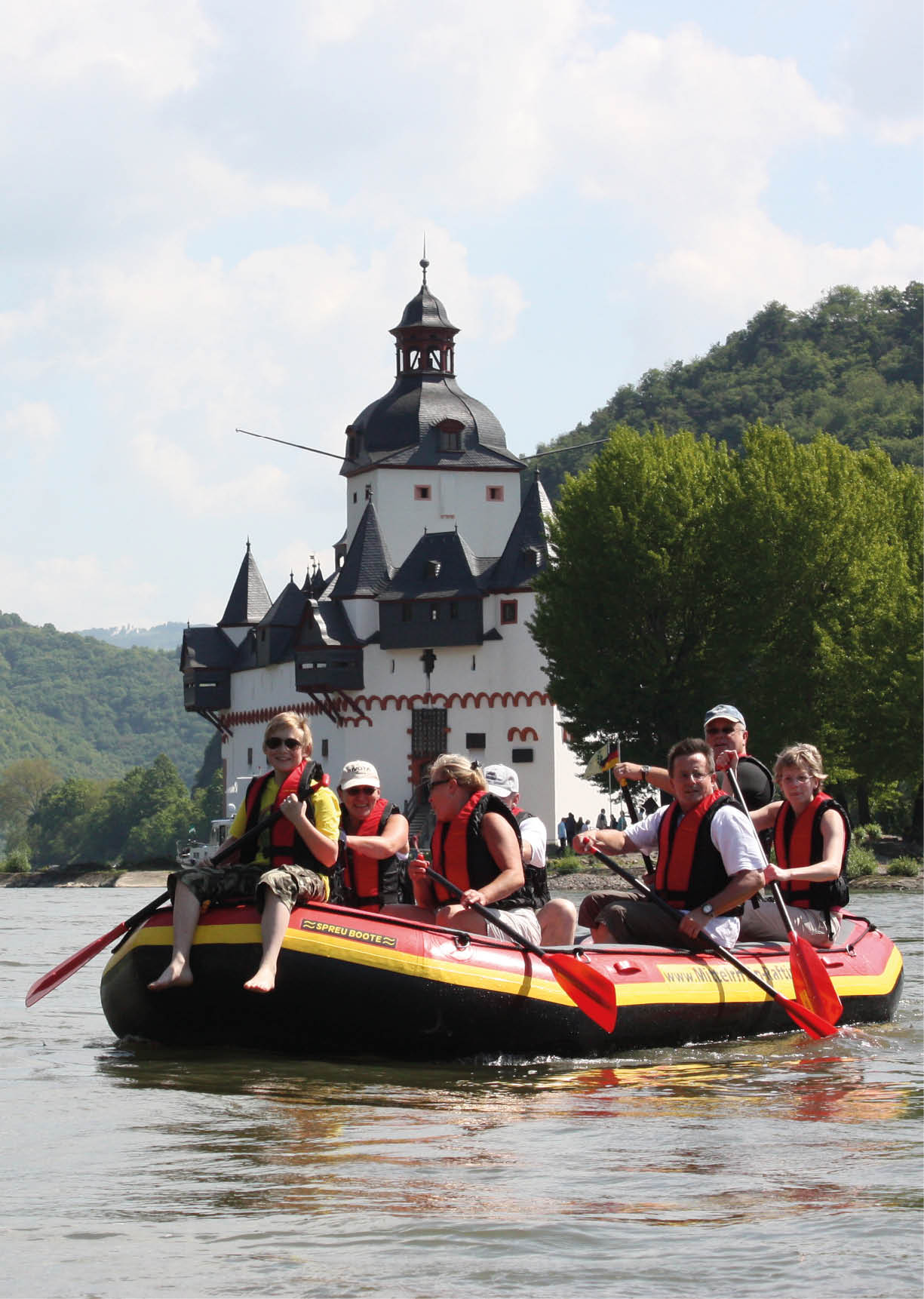 Sechs Personen mit Schwimmwesten sitzen in einem Schlauchboot auf einem Fluss vor einem historischen Schloss mit T rmen und einem bewaldeten H gel im Hintergrund. Beschreibung von KI generiert