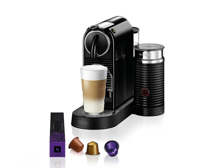 Eine schwarze Kapsel-Kaffeemaschine mit gl nzender Oberfl che steht auf einer ebenen Fl che. Daneben befindet sich ein Milchaufsch umer mit rotem Ring auf der Vorderseite. Vor der Maschine liegen drei unterschiedlich farbige Kaffeekapseln und eine l ngliche Verpackung mit sichtbarem Markenlogo. Ein hohes Glas mit mehreren Schichten Kaffee und Milchschaum steht auf der Abstellfl che der Maschine.