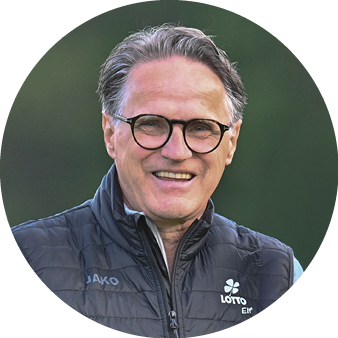 Eine Person mit grauem Haar und Brille tr gt eine dunkle, gesteppte Jacke und l chelt vor einem unscharfen gr nen Hintergrund. Beschreibung von KI generiert