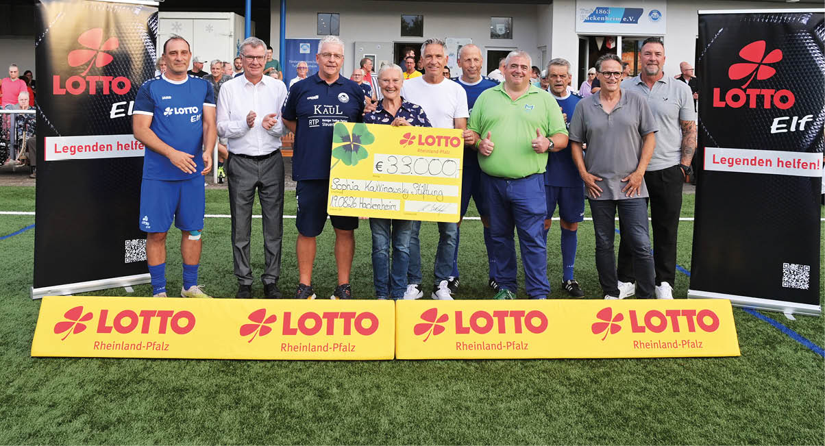 Mehrere Erwachsene stehen auf einem Sportplatz nebeneinander und halten gemeinsam einen gro en symbolischen Scheck, umgeben von Werbebannern der Lotto Rheinland-Pfalz. Beschreibung von KI generiert