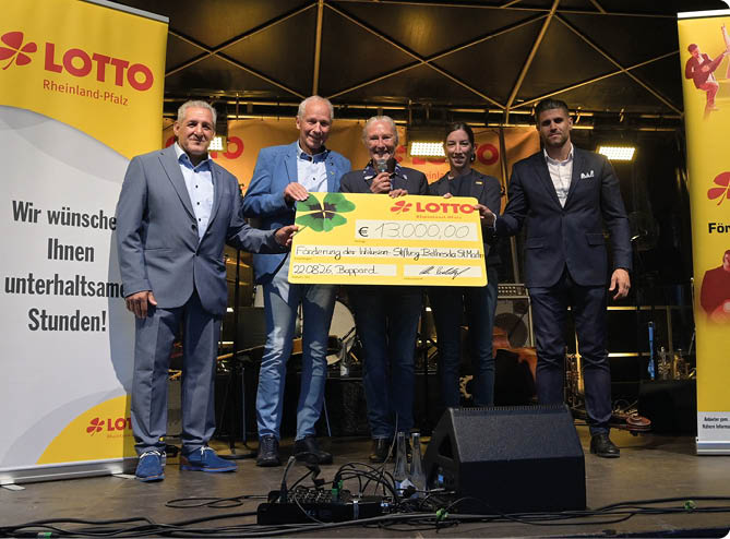 F nf Personen in formeller Kleidung stehen auf einer B hne und halten gemeinsam einen gro en symbolischen Scheck vor zwei gelben Werbebannern mit Lotto-Logos. Beschreibung von KI generiert