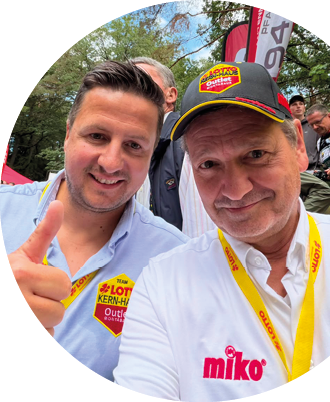 Zwei Personen stehen nah beieinander, eine tr gt eine M tze mit gelbem Rand und die andere hat kurze Haare, im Hintergrund ist ein dunkler Anzug zu sehen. Beschreibung von KI generiert