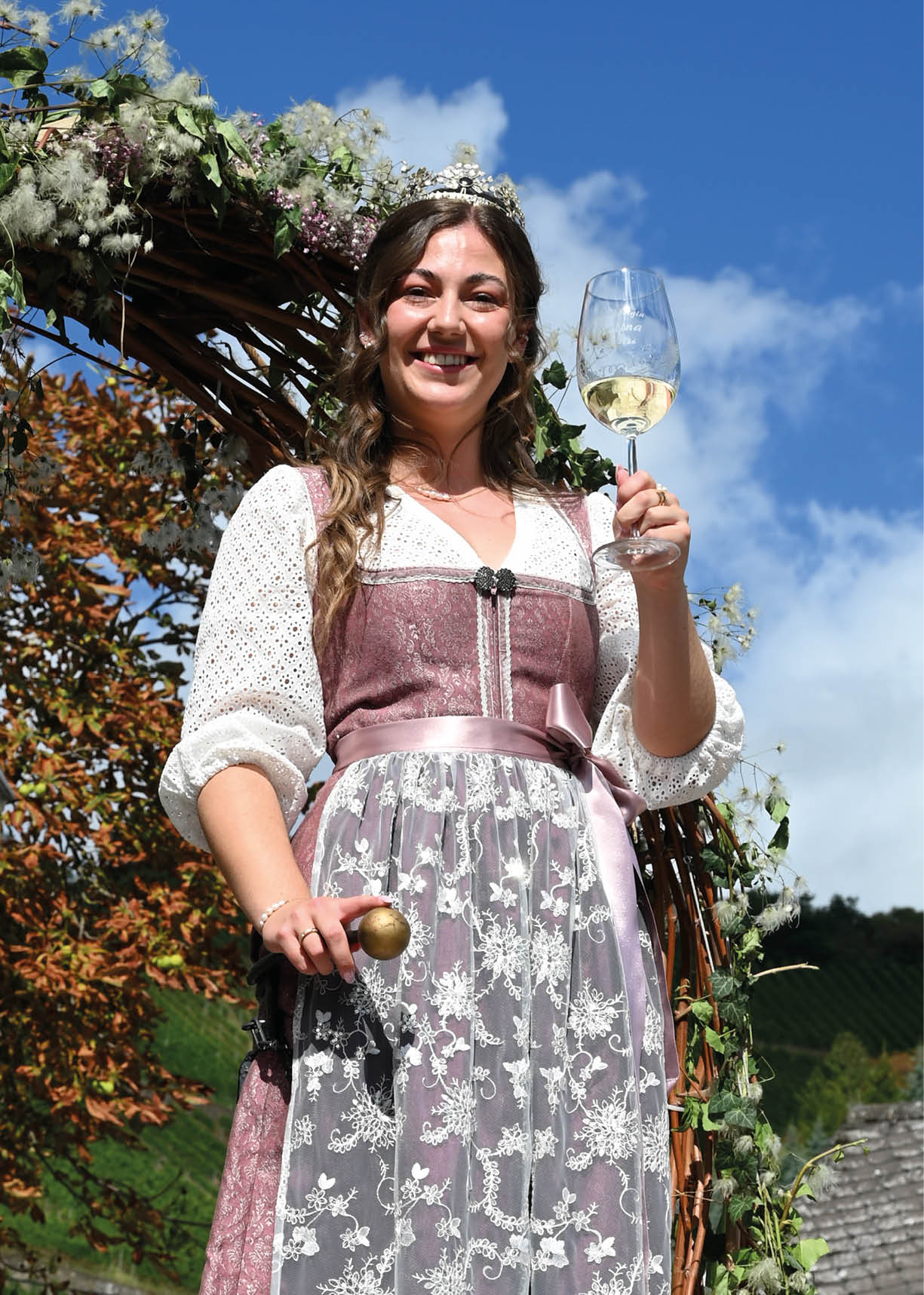Eine Person in traditioneller Tracht steht l chelnd unter einem mit Blumen geschm ckten Bogen und h lt ein Glas Wei wein in der Hand. Beschreibung von KI generiert