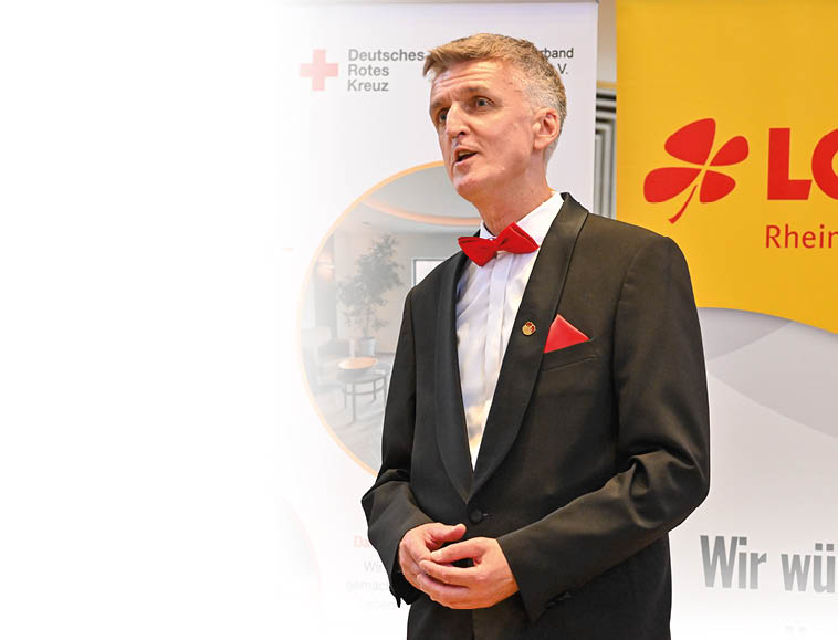 Eine Person in einem schwarzen Anzug mit roter Fliege und rotem Einstecktuch steht vor Bannern mit Logos des Deutschen Roten Kreuzes und Lotto Rheinland-Pfalz. Beschreibung von KI generiert