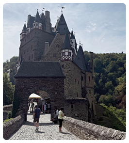Eine mittelalterliche Burg mit mehreren T rmen steht am Ende eines gepflasterten Weges, auf dem mehrere Personen gehen, umgeben von bewaldeten H geln unter einem bew lkten Himmel. Beschreibung von KI generiert