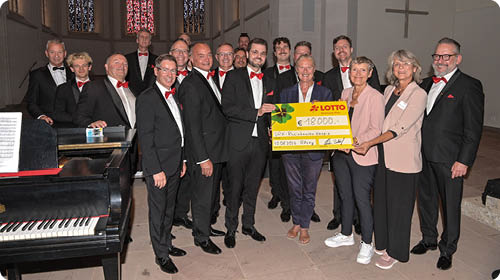 Eine Gruppe von Erwachsenen in formeller Kleidung steht in einem Kirchenraum zusammen und h lt gemeinsam einen gro en symbolischen Scheck. Beschreibung von KI generiert