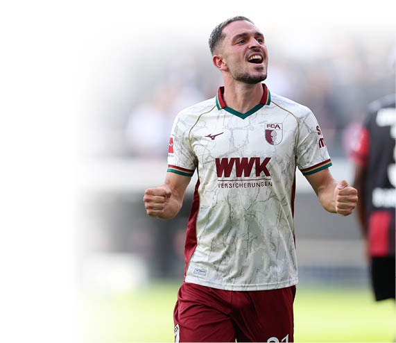 Ein jubelnder Spieler in wei -rotem Fu balltrikot steht auf dem Spielfeld, w hrend eine weitere Person in schwarzem Trikot mit der Nummer drei im Hintergrund zu sehen ist. Beschreibung von KI generiert
