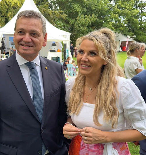 Zwei Erwachsene in eleganter Kleidung stehen l chelnd im Freien vor wei en Zelten und gr nen B umen bei einer Veranstaltung. Beschreibung von KI generiert