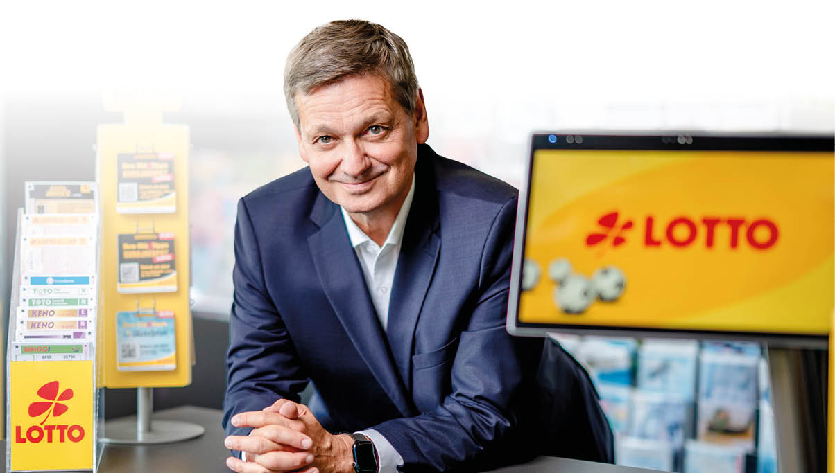 Eine Person in einem Anzug sitzt l chelnd an einem Tisch mit Lotto-Werbematerialien und einem Bildschirm mit dem Lotto-Logo im Hintergrund. Beschreibung von KI generiert