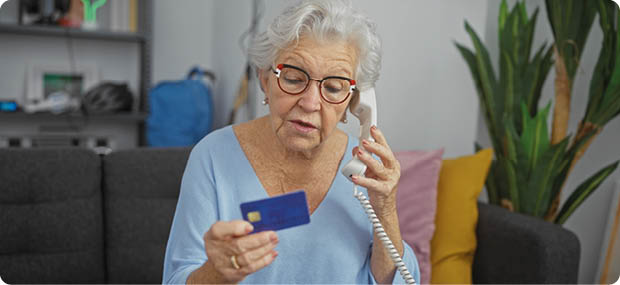 Senior mit Brille und hellblauem Pullover sitzt auf einem Sofa und h lt eine Telefonh rer in der einen und eine Karte in der anderen Hand. Beschreibung von KI generiert