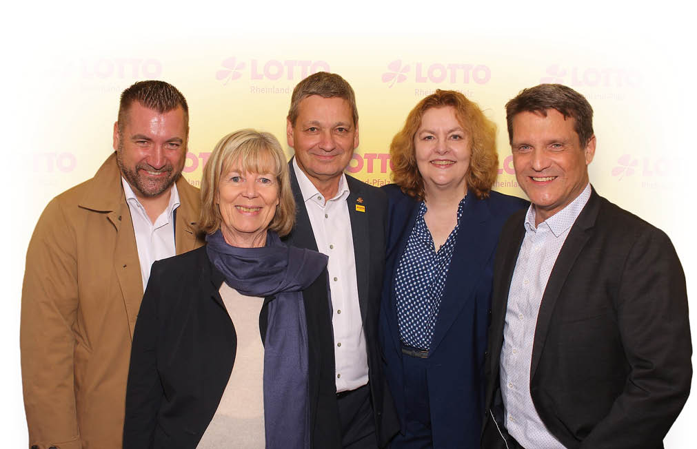 F nf Erwachsene stehen eng beieinander und l cheln vor einem hellen Hintergrund mit dem Schriftzug Lotto. Beschreibung von KI generiert