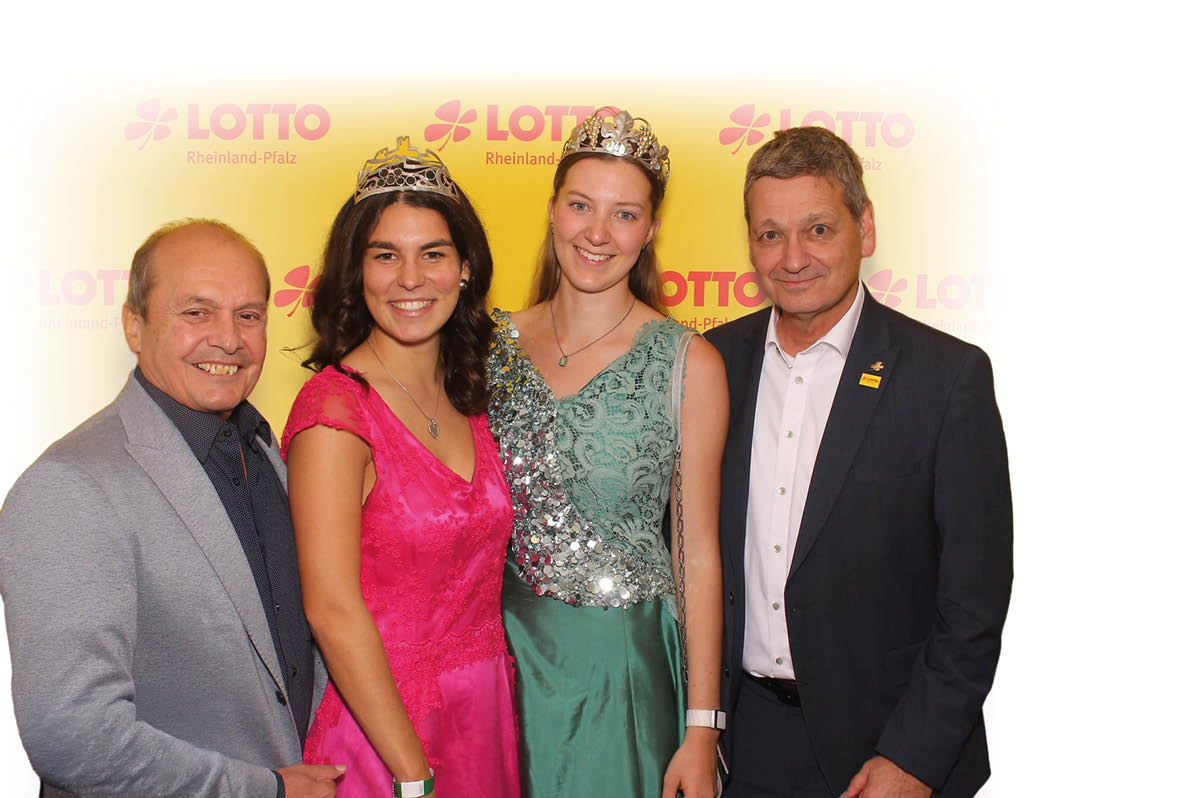 Vier Personen stehen vor einem gelben Hintergrund mit Lotto-Logo, zwei davon tragen Kronen und festliche Kleider, w hrend die anderen beiden Anz ge tragen und alle l cheln. Beschreibung von KI generiert
