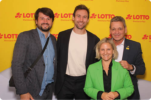 Vier Erwachsene stehen vor einer gelben Wand mit Lotto-Logo, drei von ihnen tragen Anz ge und eine tr gt ein gr nes Sakko, alle l cheln in die Kamera. Beschreibung von KI generiert