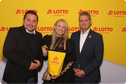 Drei Erwachsene stehen vor einer gelben Wand mit Lotto-Logo, wobei eine Person in der Mitte eine gelbe Lotteriet te h lt und alle l cheln. Beschreibung von KI generiert