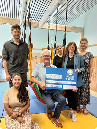 Sechs Personen in einem hellen Raum mit Turnmatten und Schlingen halten gemeinsam einen gro en symbolischen Scheck mit dem Logo von Gl cksSpirale und l cheln in die Kamera. Beschreibung von KI generiert