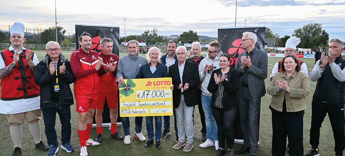 Eine Gruppe von Menschen steht auf einem Sportplatz und h lt gemeinsam einen gro en symbolischen Scheck, w hrend sie applaudieren. Beschreibung von KI generiert