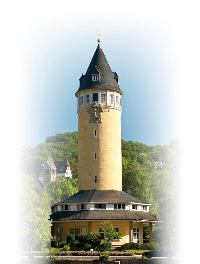 Ein hoher, runder Turm mit spitzem Dach steht an einem Flussufer vor bewaldeten H geln und mehreren H usern. Beschreibung von KI generiert