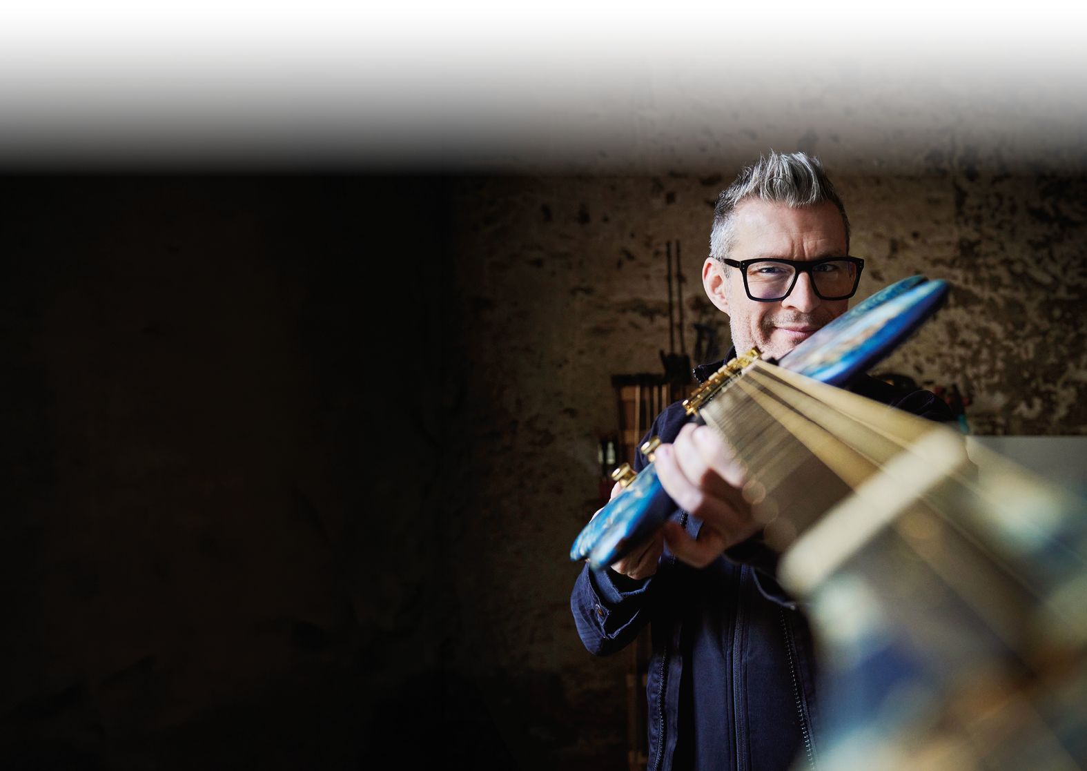 Eine Person mit Brille und grauen Haaren spielt l chelnd eine Gitarre in einem dunklen Raum. Beschreibung von KI generiert