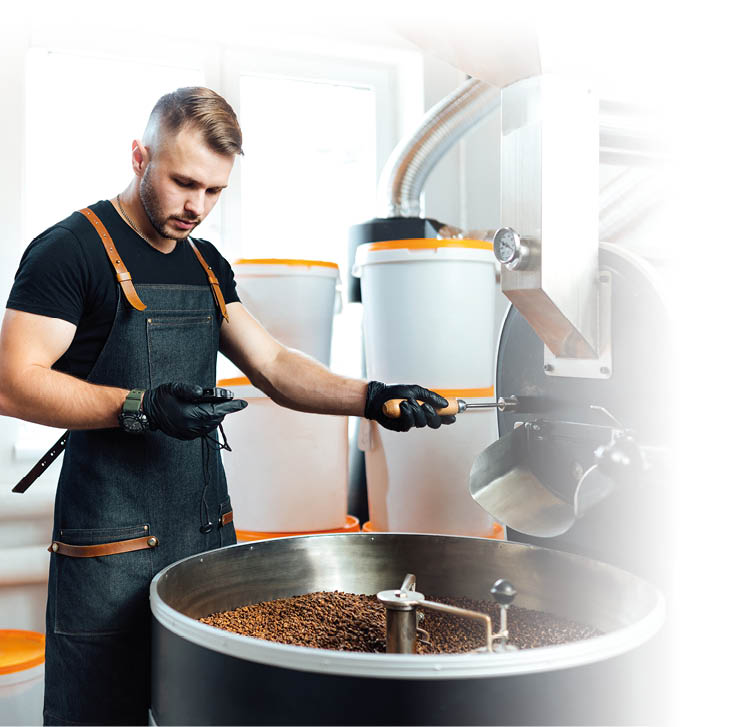 Eine Person mit Sch rze und Handschuhen berpr ft ger stete Kaffeebohnen in einer industriellen R stmaschine in einem hellen Raum.  Beschreibung von KI generiert
