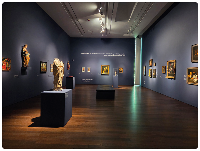 Ein Museumssaal mit dunklen W nden zeigt verschiedene Gem lde und Skulpturen, die auf Sockeln und an den W nden ausgestellt sind, beleuchtet von Deckenstrahlern. Beschreibung von KI generiert