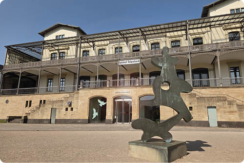 Vor einem historischen Geb ude mit Balkon und mehreren Fenstern steht eine abstrakte Skulptur aus Metall auf einem Sockel. Beschreibung von KI generiert