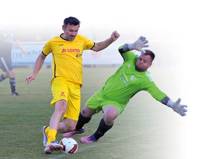 Zwei Personen in Sportkleidung k mpfen auf einem Fu ballfeld um den Ball, wobei eine in gelber Kleidung steht und die andere in gr ner Torwartkleidung auf dem Boden rutscht. Beschreibung von KI generiert