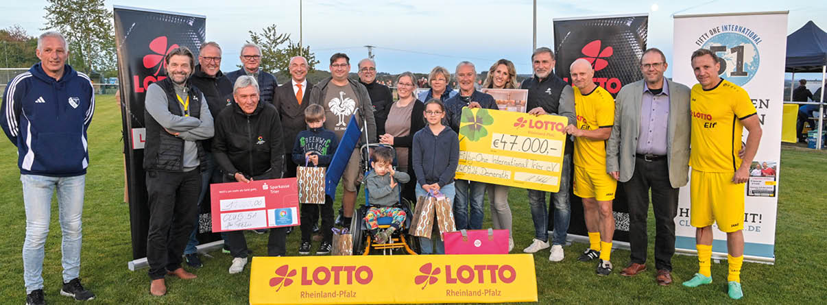 Eine Gruppe von Menschen steht auf einem Rasen bei Tageslicht zusammen, einige halten Geschenke und einen gro en symbolischen Scheck, w hrend zwei Personen in gelber Sportkleidung posieren. Beschreibung von KI generiert