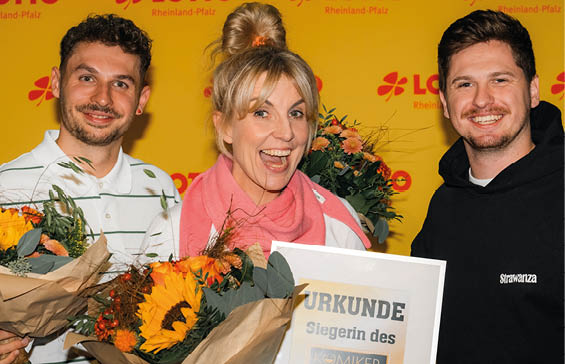 Drei Personen mit Blumenstr u en und einer Urkunde l cheln vor einer gelben Wand mit Lotto Rheinland-Pfalz-Logo. Beschreibung von KI generiert