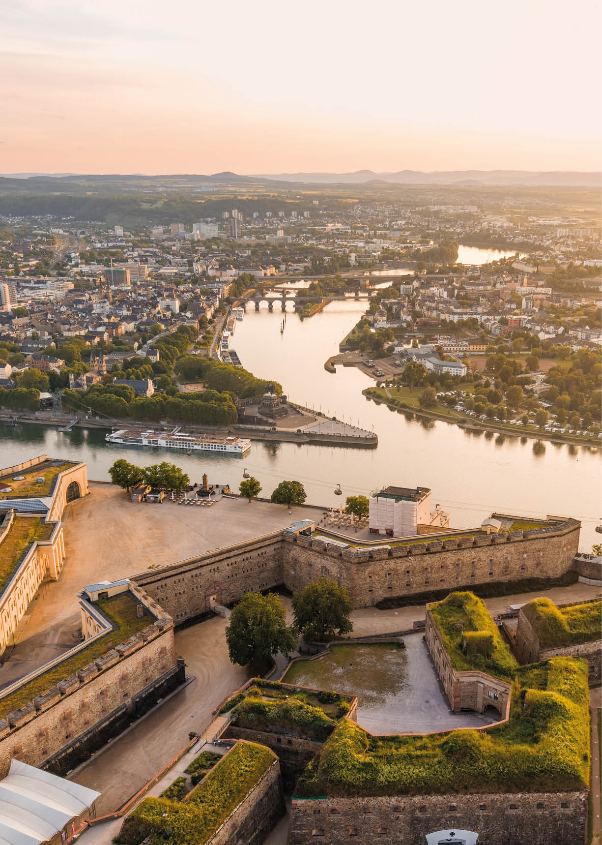 Eine historische Festungsanlage mit gr nen Grasd chern liegt an einem Fluss, der sich durch eine Stadt mit vielen Geb uden und Br cken im Hintergrund schl ngelt. Beschreibung von KI generiert