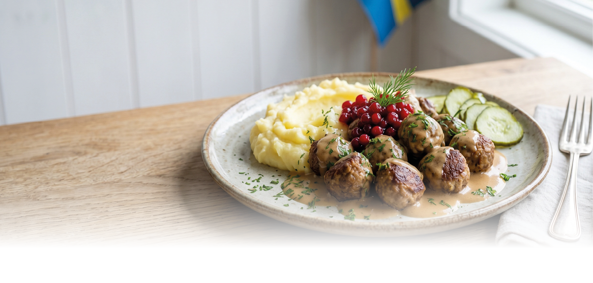 Auf einem Holztisch steht ein Teller mit schwedischen Fleischb llchen, Kartoffelp ree, Gurkenscheiben und Preiselbeeren neben Besteck auf einer Serviette, im Hintergrund ist eine schwedische Flagge zu sehen. Beschreibung von KI generiert