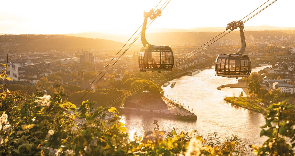 Zwei Gondeln einer Seilbahn schweben ber einem Fluss mit gr ner Ufervegetation und einer Stadt im Hintergrund bei Sonnenuntergang.  Beschreibung von KI generiert