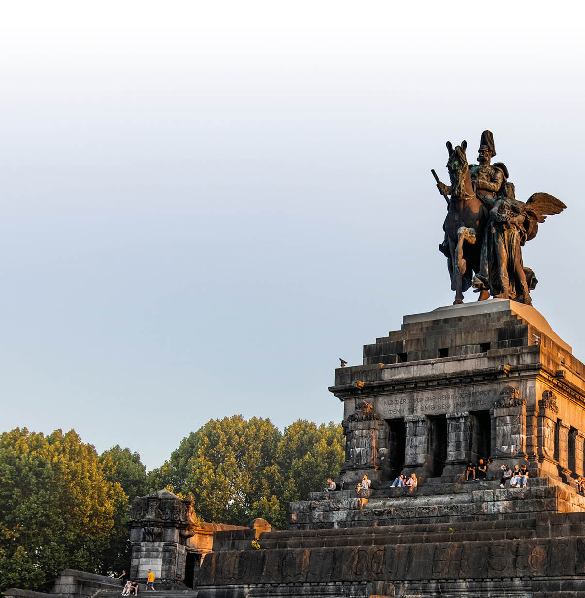 Ein gro es steinernes Denkmal mit einer Reiterstatue und einer gefl gelten Figur steht auf einer erh hten Plattform vor einem Hintergrund aus B umen und klarem Himmel. Beschreibung von KI generiert