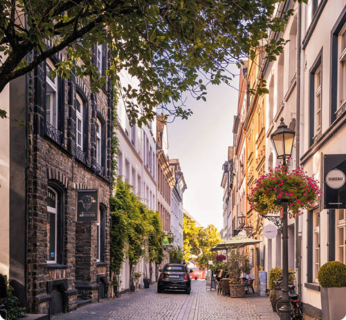 Eine gepflasterte Stra e mit parkenden Autos und Fahrr dern wird von Geb uden mit Blumen und B umen an den Seiten ges umt, w hrend Menschen an einem Stra encaf sitzen.  Beschreibung von KI generiert