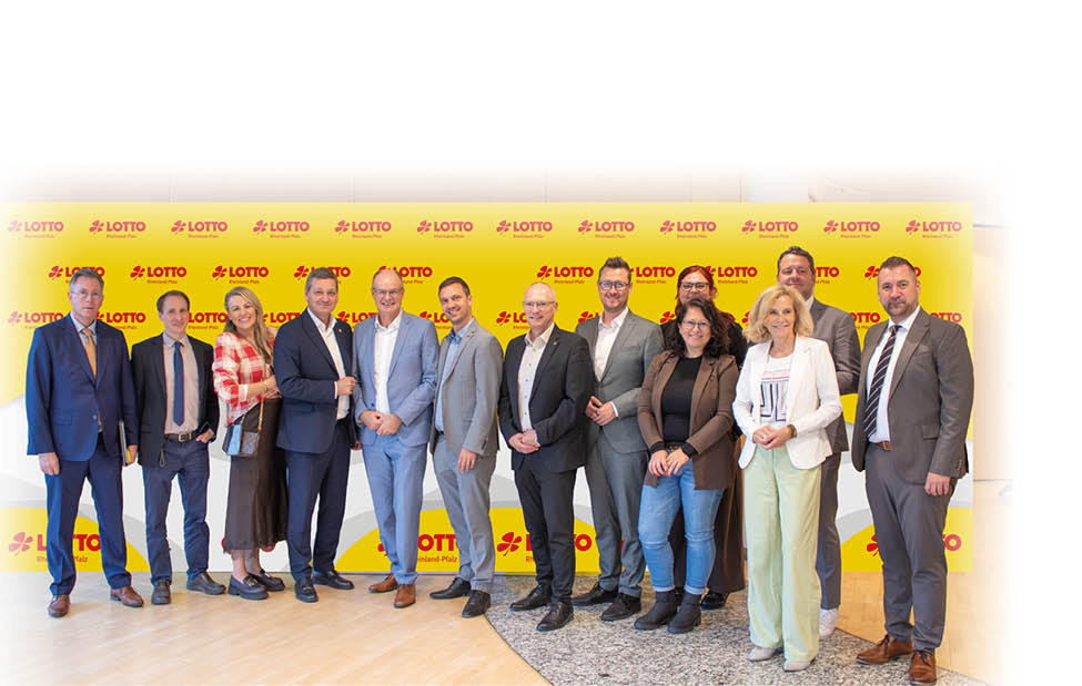 Eine Gruppe von Erwachsenen in formeller und halbformeller Kleidung steht vor einer gelben Wand mit dem Logo “LOTTO" und posiert f r ein Gruppenfoto. Beschreibung von KI generiert