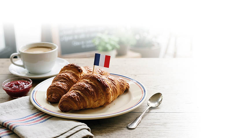 Zwei Croissants mit einer kleinen franz sischen Flagge auf einem Teller stehen neben einer Tasse Kaffee, einem L ffel und einem kleinen Sch lchen Marmelade auf einem Holztisch. Beschreibung von KI generiert