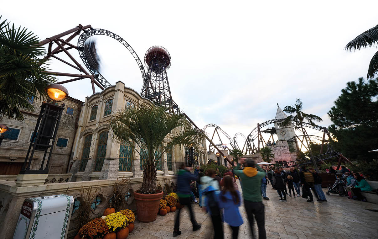 Mehrere Menschen gehen auf einem gepflasterten Weg an einem Freizeitpark mit einer Achterbahn und herbstlicher Dekoration aus K rbissen und Blumen vorbei. Beschreibung von KI generiert