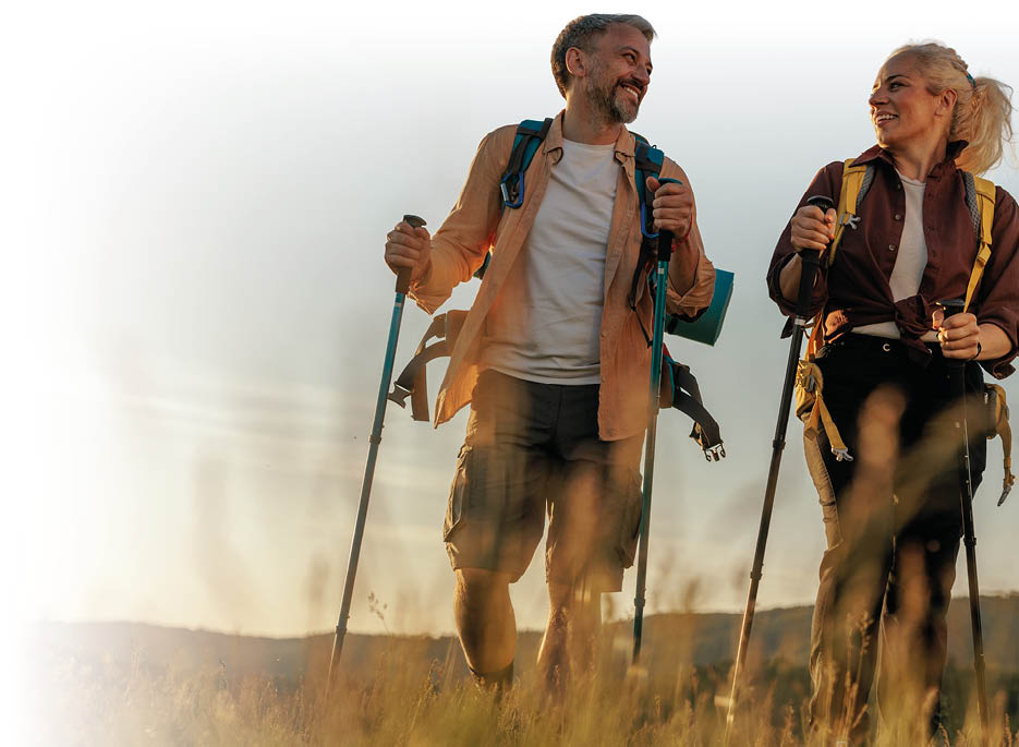 Zwei Erwachsene mit Wanderst cken und Rucks cken gehen l chelnd durch ein hohes Grasfeld bei Sonnenuntergang. Beschreibung von KI generiert