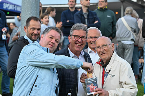 F nf Erwachsene stehen drau en auf einer Veranstaltung zusammen und l cheln in die Kamera, wobei einer eine Karte oder ein Ticket h lt. Beschreibung von KI generiert