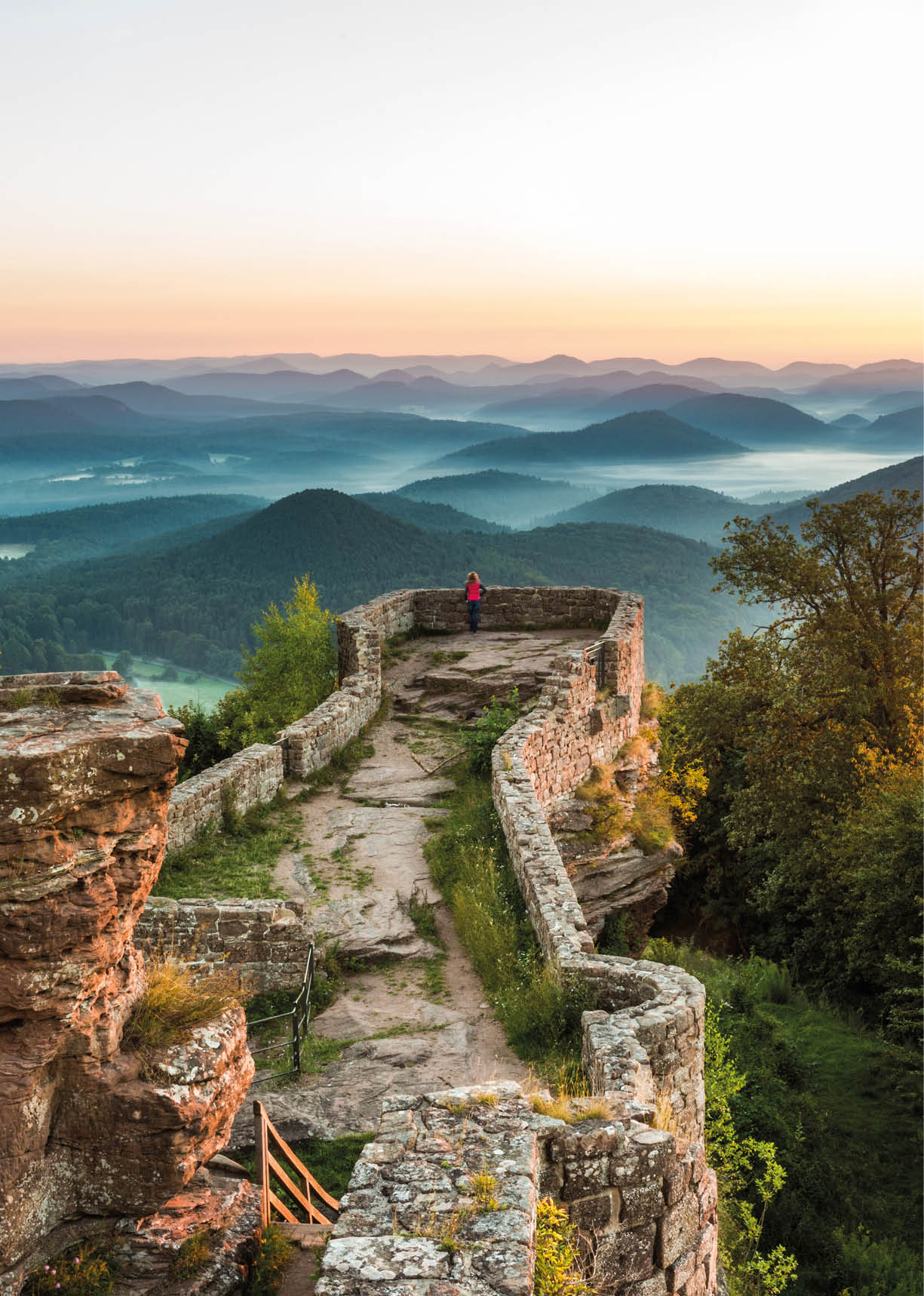 Eine Person in roter Kleidung steht auf einer alten Steinmauer mit Blick auf eine neblige Berglandschaft bei Sonnenaufgang oder Sonnenuntergang. Beschreibung von KI generiert