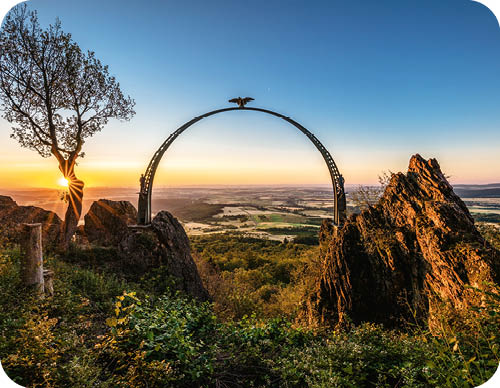 Eine metallene Torbogenstruktur mit einem Adler darauf steht auf einem bewachsenen H gel mit Felsen und einem Baum im Sonnenuntergang ber einer weiten Landschaft.  Beschreibung von KI generiert