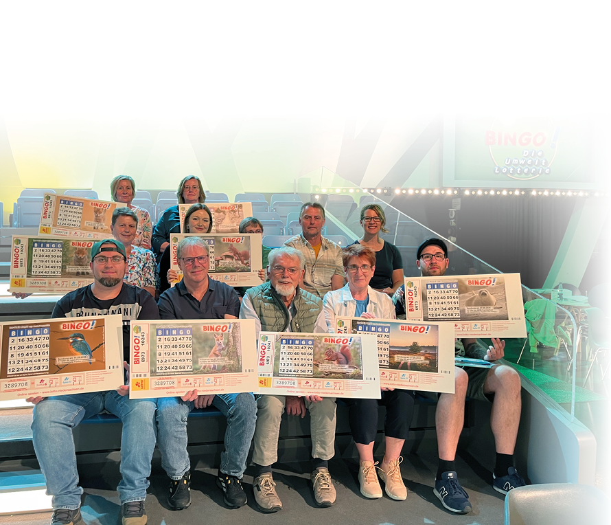 Eine Gruppe von Erwachsenen sitzt auf Trib nenstufen und h lt gro e Bingo-Karten in einer hell beleuchteten Halle mit blauem Hintergrund. Beschreibung von KI generiert